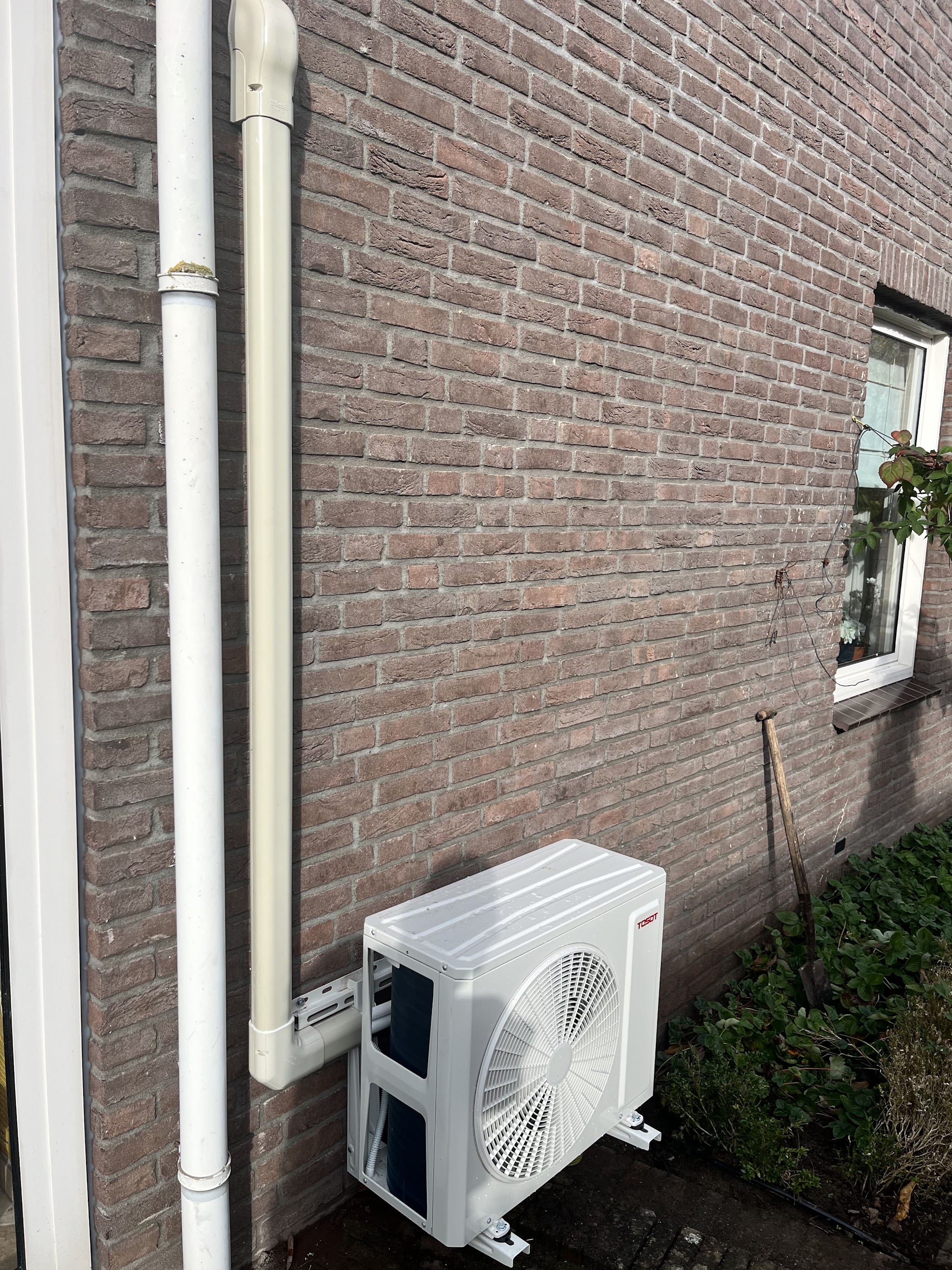Airco buitenunit op een muurbeugel naast een regenpijp aan een bakstenen gevel